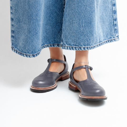 A person wearing wide-leg blue denim jeans and gray-blue Mary Jane-style leather shoes with brown soles, by Mafalda handmade, shown from the calves down on a clean white studio background.