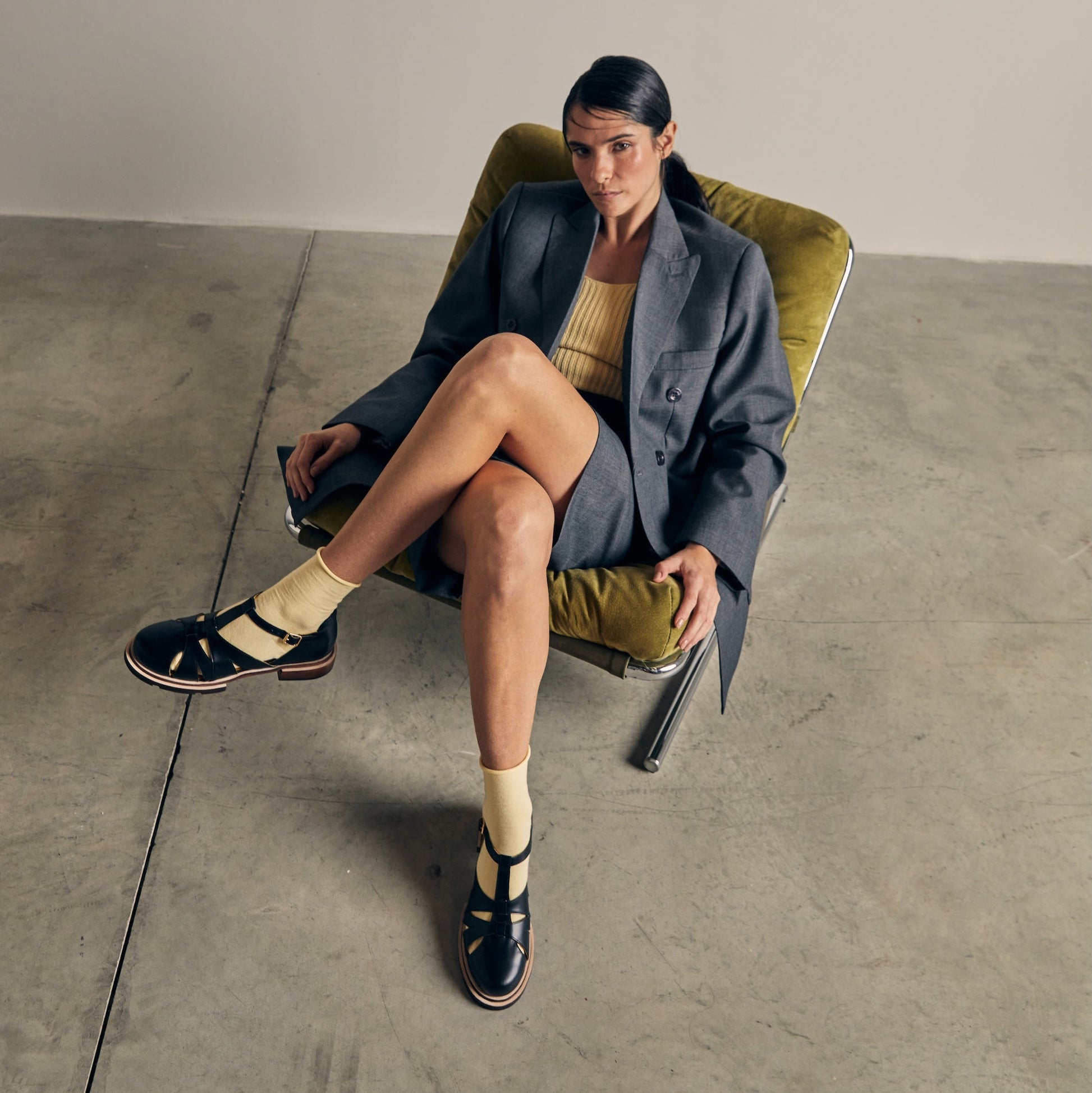 Maite Black Fisherman Sandals by Mafalda Handmade Model wearing with yellow socks and yellow top with blue blazer and skirt sat on a green chair