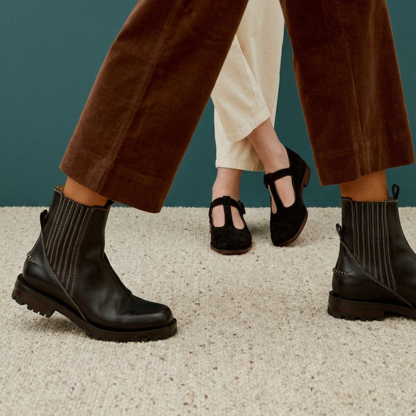 A close-up lifestyle shot of a person wearing black leather ankle boots with vertical ribbed side panels and chunky lug soles, paired with brown corduroy trousers and a dark teal background.