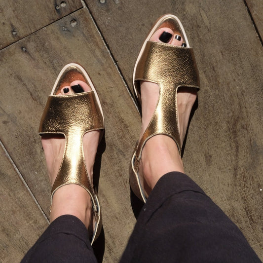 A pair of Yoko Gold Sandals, genuine leather sandals in gold color with an adjustable ankle strap and a flat sole by Mafalda Handmade worn by model with black trousers